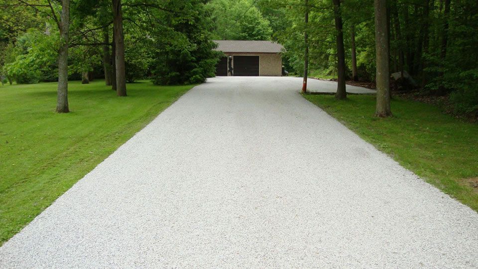 White gravel driveway bordered by green grass and trees, leading to a garage or shed in the distance