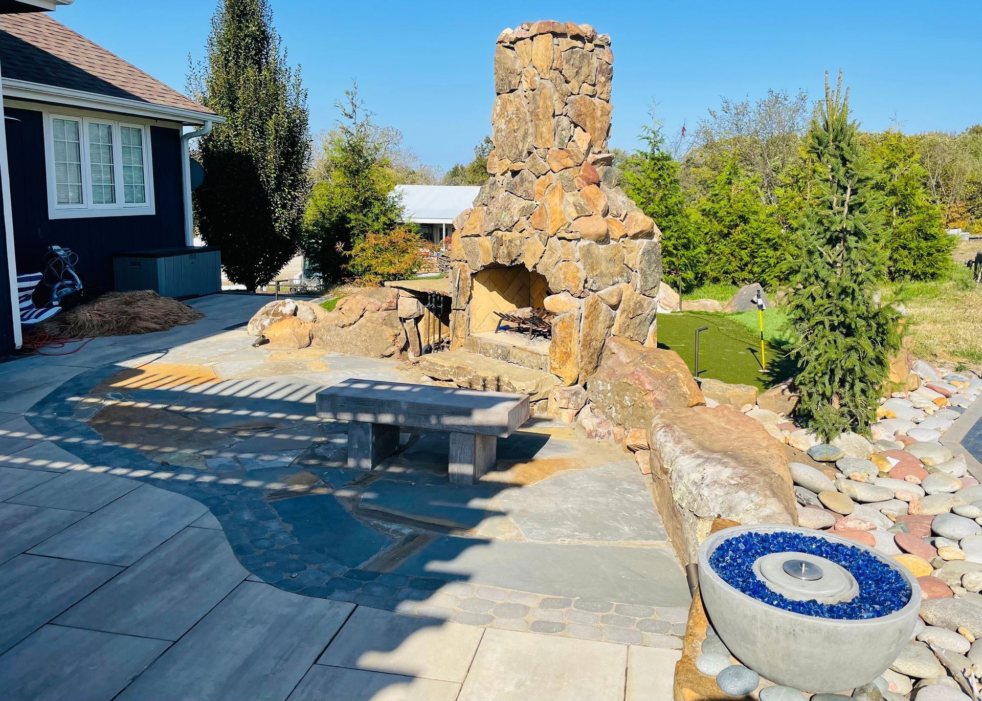 Stone outdoor fireplace beside a paved patio with a pool and landscaped garden
