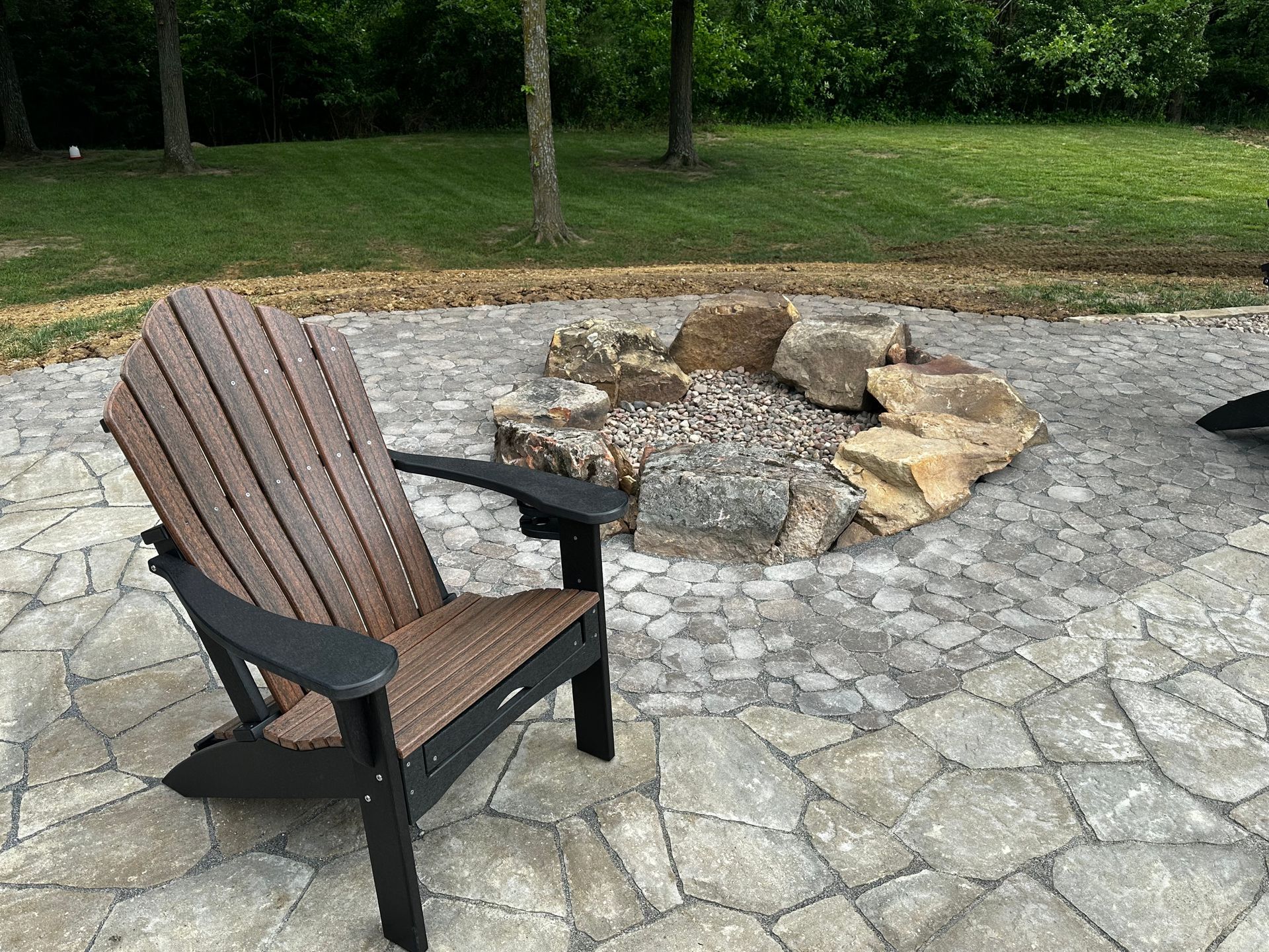 Wooden chair beside a stone fire pit on a paved patio with grass and trees in the background