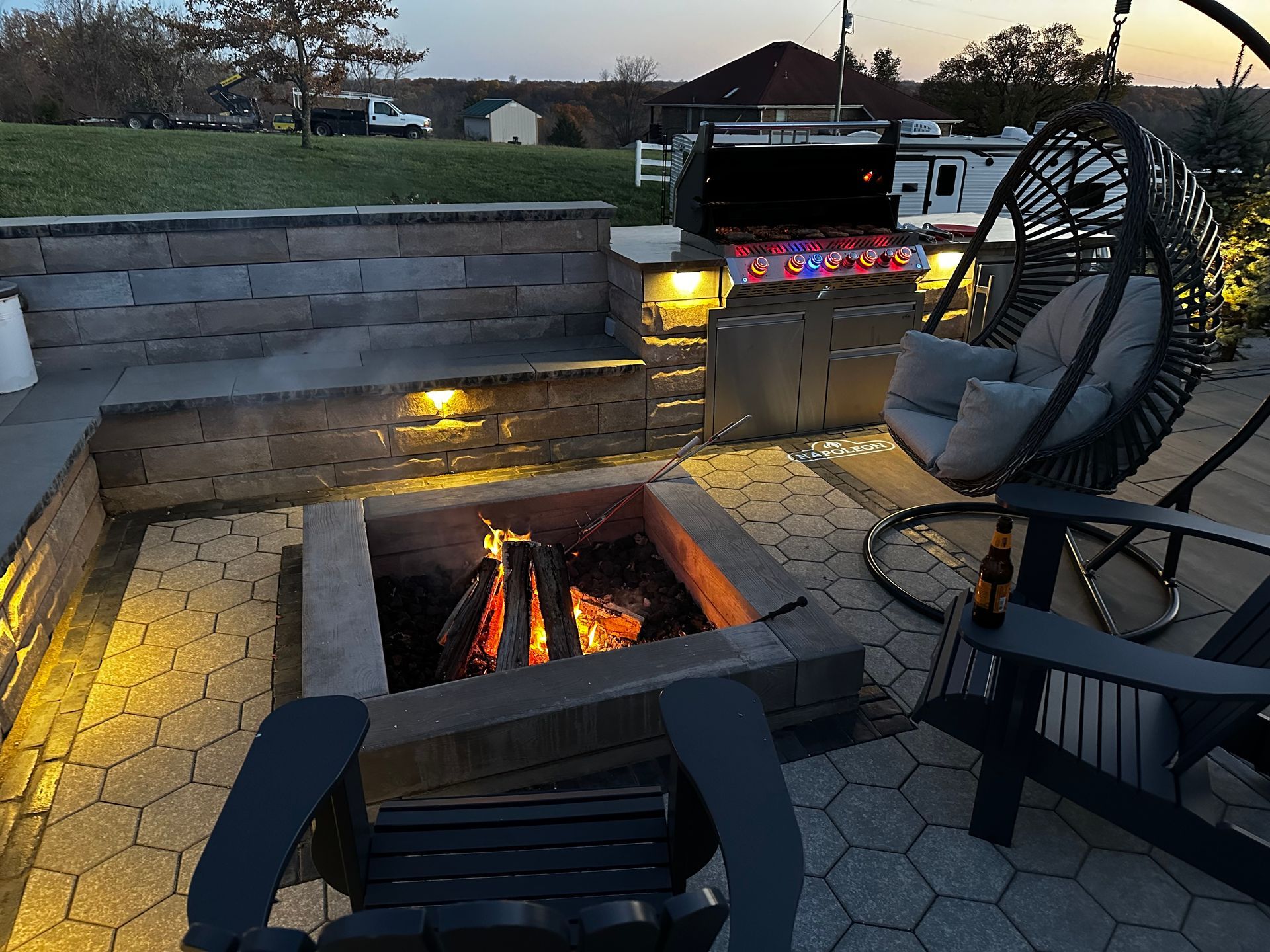 Backyard patio with a glowing fire pit, stone seating, string lights, and a hanging chair at dusk