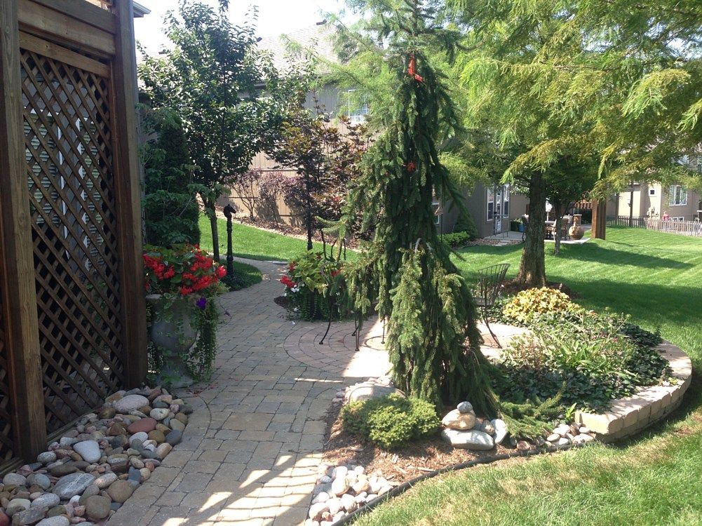 Brick pathway through landscaped backyard with green trees, flowerbeds, and a lattice fence.