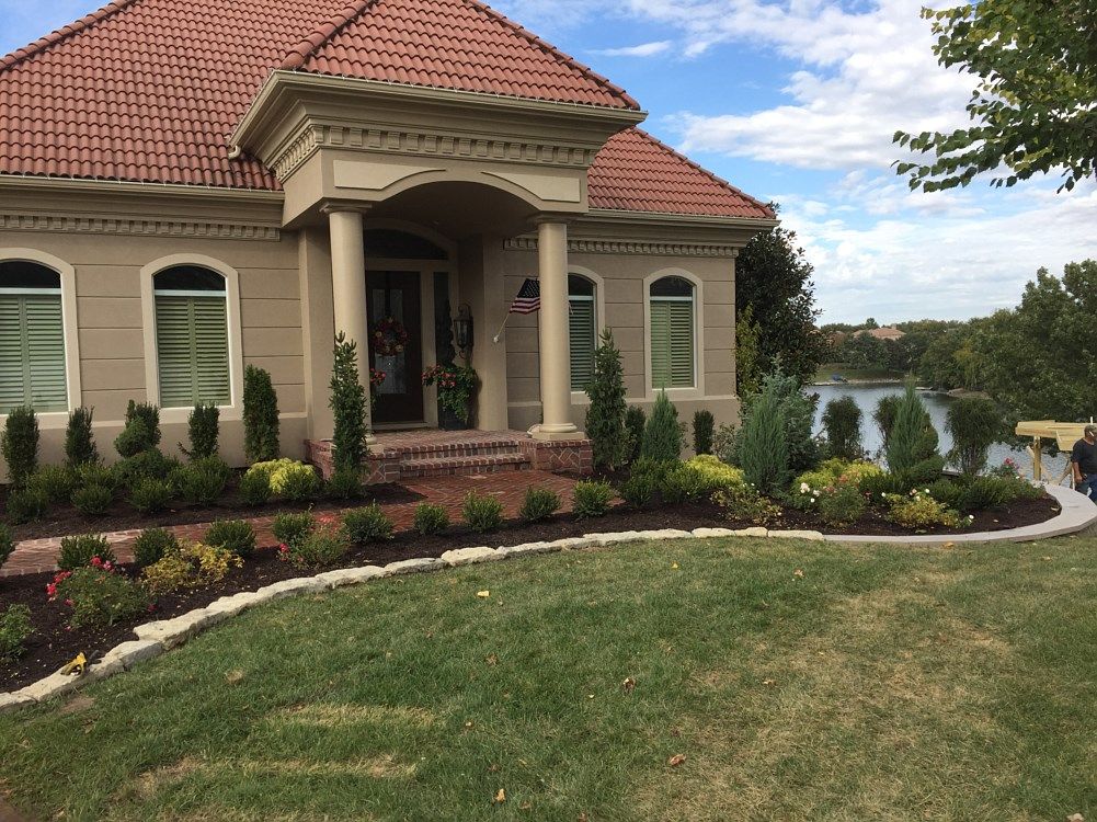 Beige house with red tile roof, columns, arched windows, and landscaped front yard next to a lake.
