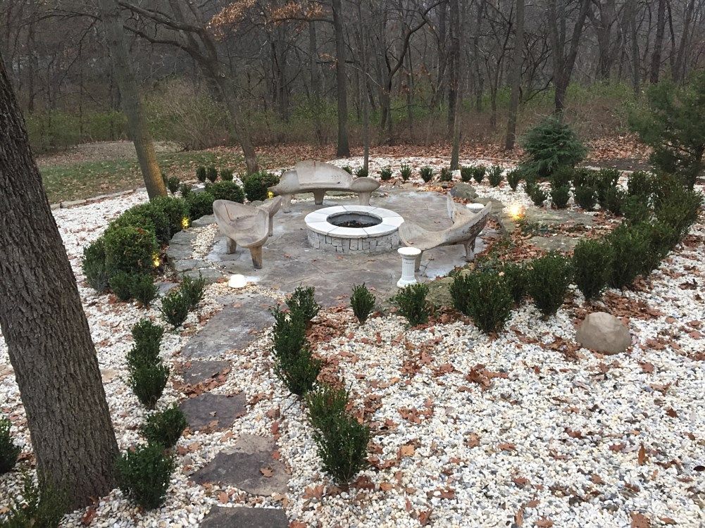 Stone fire pit surrounded by stone seating and landscaping with shrubs and white gravel in a wooded area.