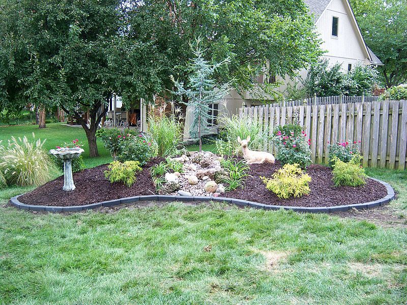 A landscaped garden bed with a small waterfall, birdbath, and wooden fence.