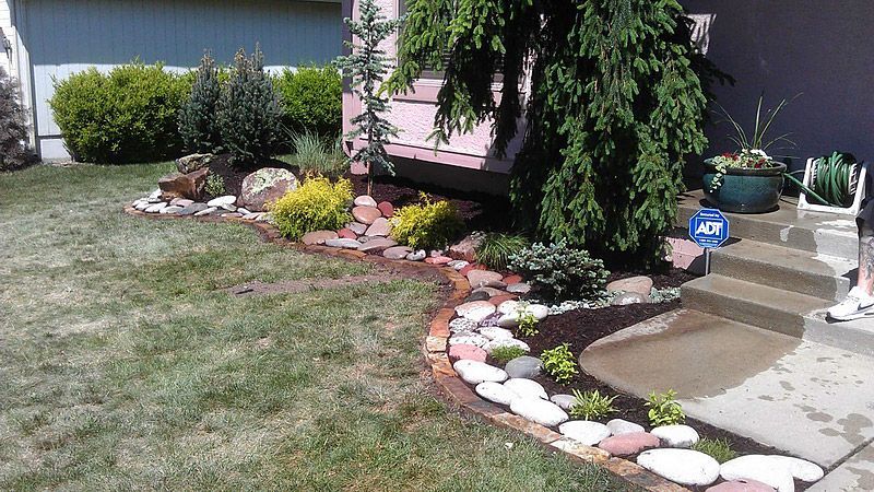 Landscaped yard with stone borders and various plants near a house and steps.