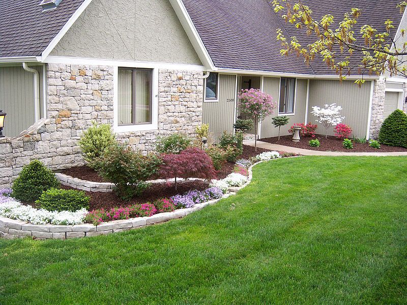 House with tiered landscaping, lush green lawn, various flowering plants and shrubs.