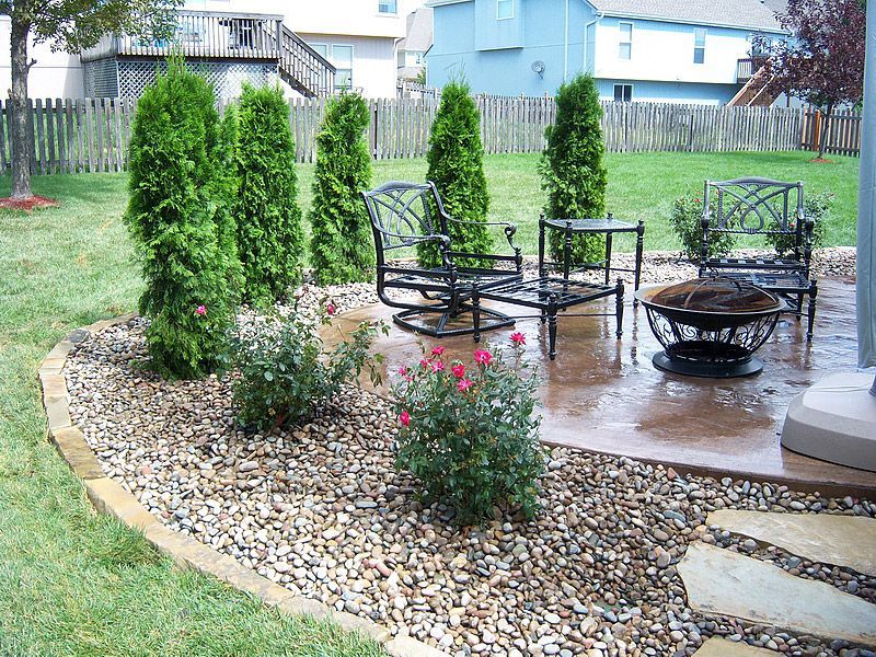 Patio with outdoor seating, fire pit, and evergreen trees surrounded by rocks in a backyard.