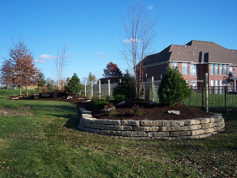 Stone-walled garden bed with trees, shrubs, and mulch on a grassy lawn in front of a brick house under a blue sky.