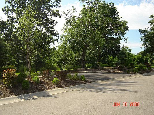 Asphalt road curves around landscaped area with trees and shrubs under a sunny sky.
