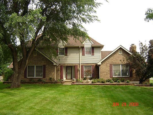 Two-story brick house with brown roof and burgundy shutters, in front of a green lawn and large tree.