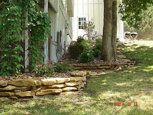 Stone-edged garden bed with flowers and a tree, next to a white building. Green grass and foliage.