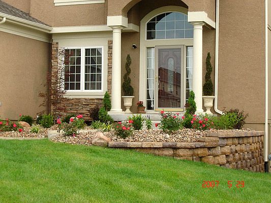 Tan house with arched doorway, pillars, and flowerbeds with roses.