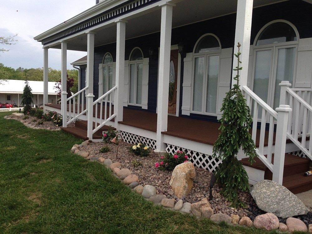 A blue house with a white porch and flowerbed landscaping, under a cloudy sky.