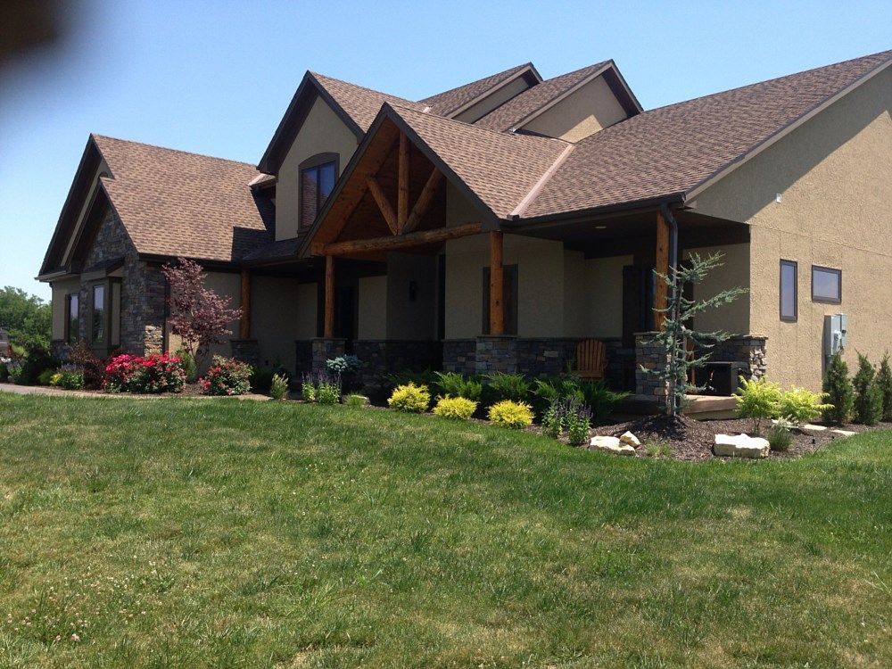 Beige stucco house with a brown roof and wooden porch, set on a green lawn with landscaping under a blue sky.