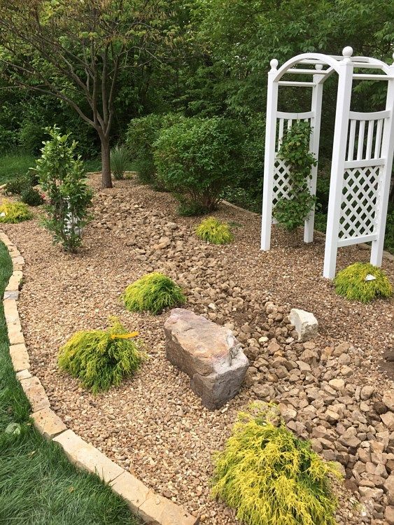 A garden bed with tan gravel, rocks, and green and yellow plants, including an arbor with a trellis.