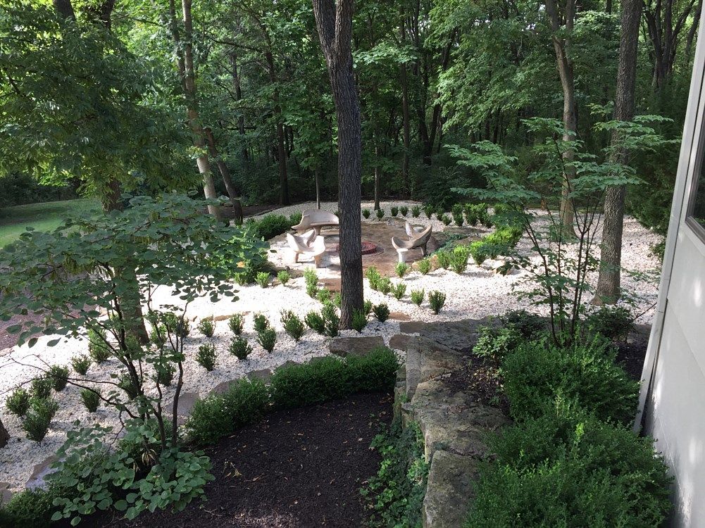 A landscaped backyard with white gravel, small plants, and a picnic table in a wooded setting.