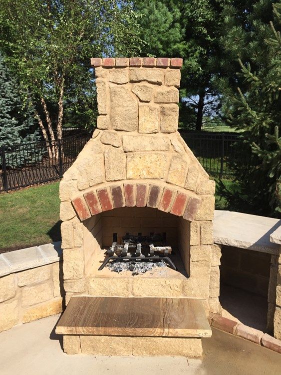 Stone outdoor fireplace with brick details and metal grate, on a patio surrounded by trees.