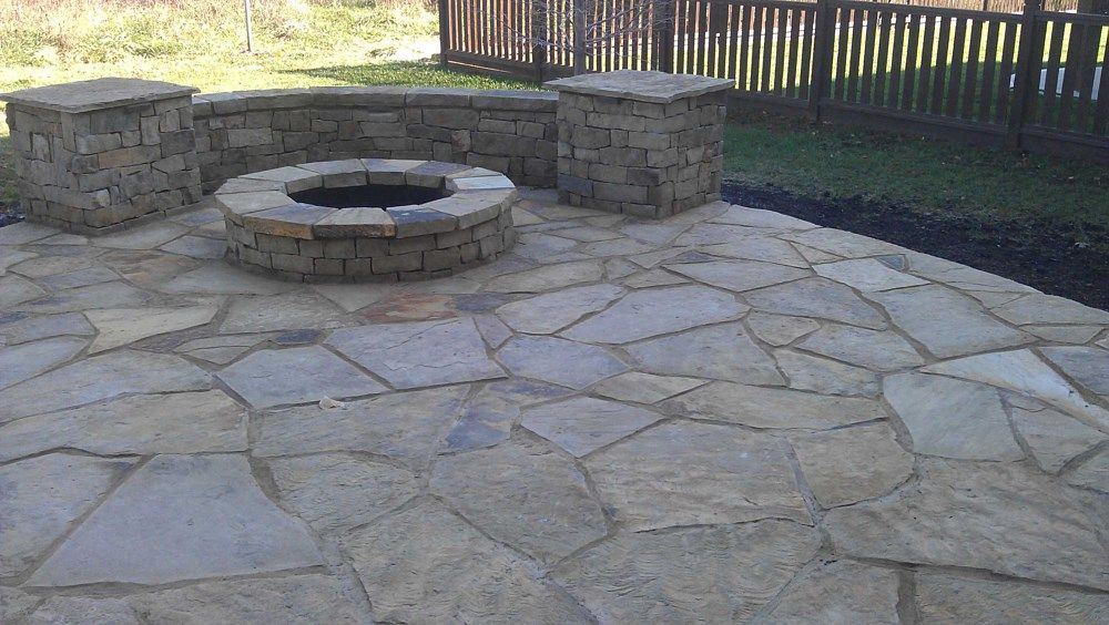 Stone patio with fire pit, seating wall, and pillars next to a wooden fence.