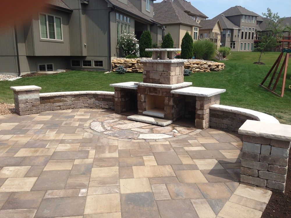Stone patio with built-in fireplace and seating area, surrounded by green grass and upscale homes.