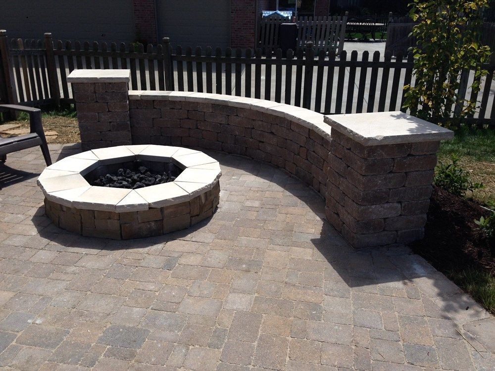 Stone fire pit and curved seating on a paved patio, black picket fence in background.