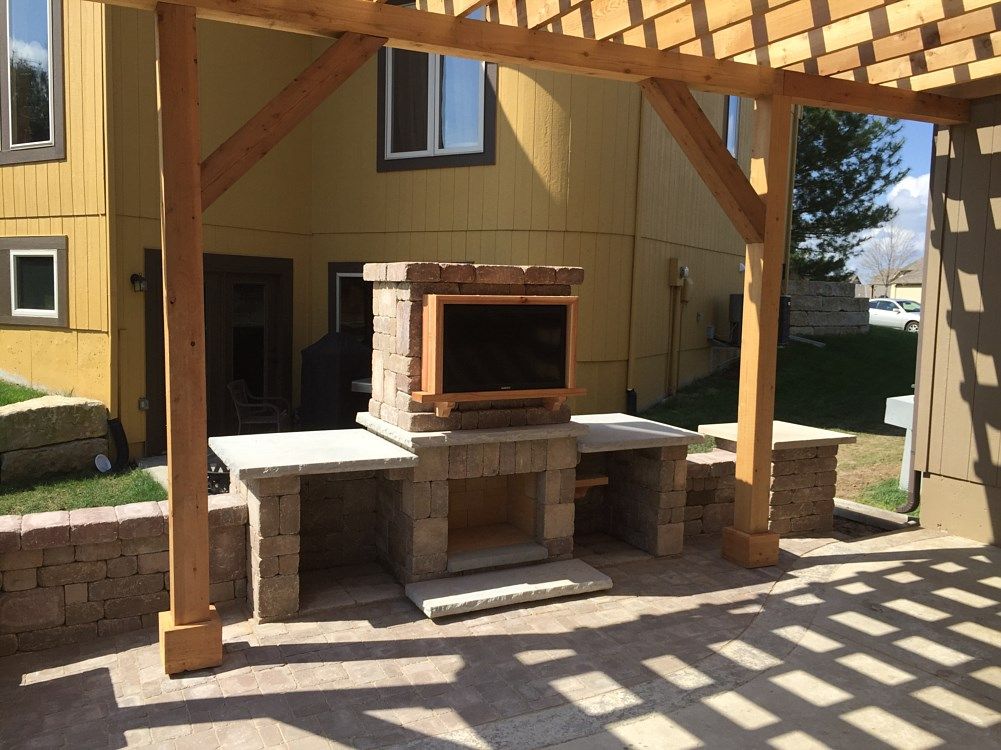 Outdoor kitchen under a wooden pergola with a stone fireplace, counters, and a TV.