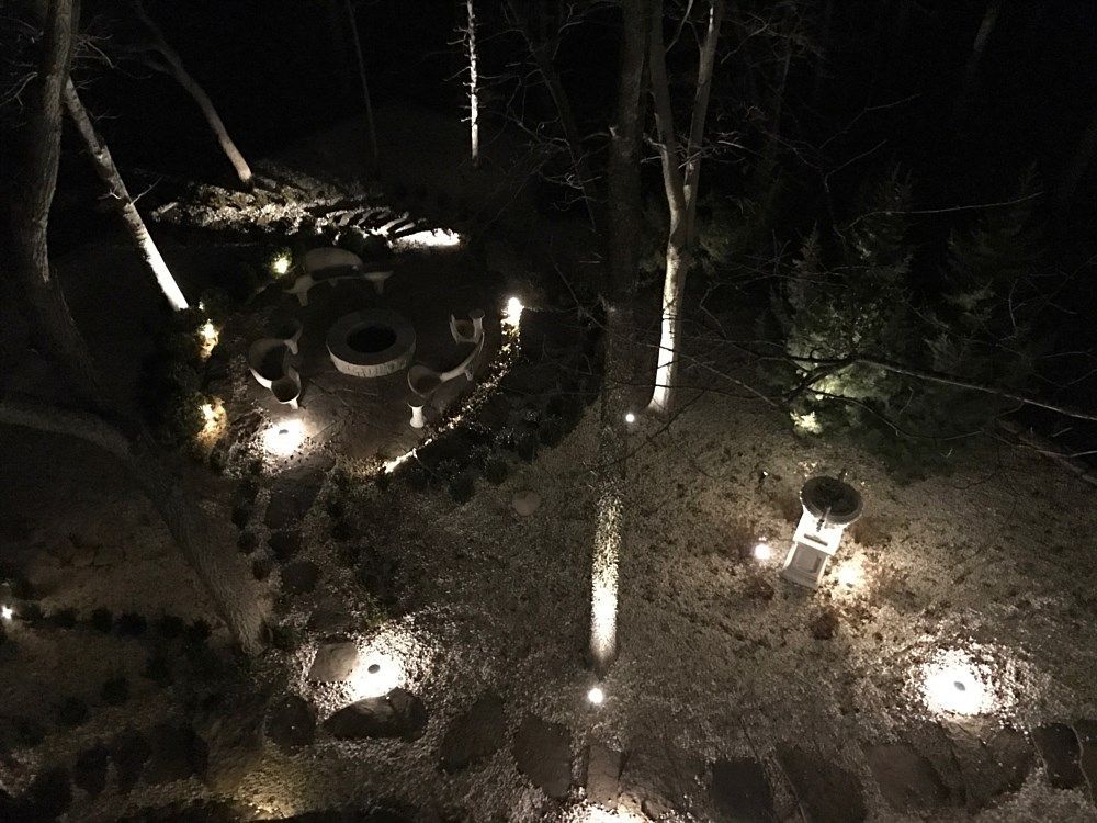 Nighttime garden with ground lights illuminating trees, a fire pit area, and a stone pathway.