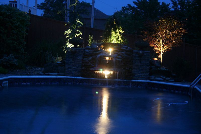 Nighttime view of a pool with a lit waterfall feature; lights reflect on the water.