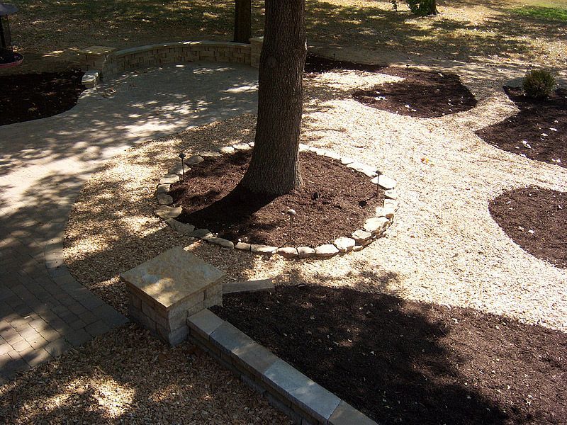 Gravel pathways and mulch beds surround a tree trunk in a sunny yard with stone borders and a low wall.