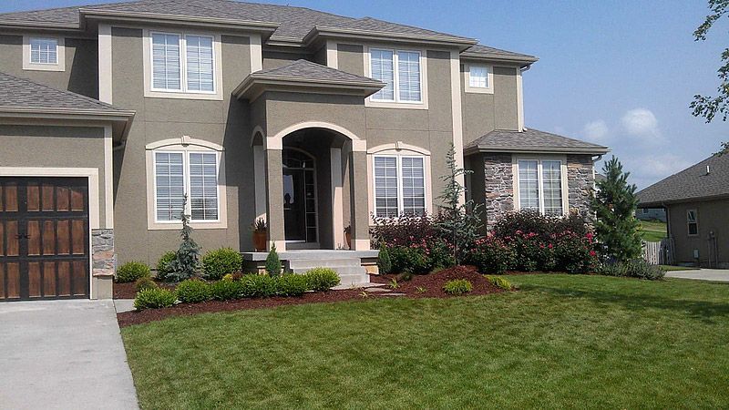 Two-story house with green lawn, landscaping, and tan stucco exterior. Dark brown garage door and gray roof.