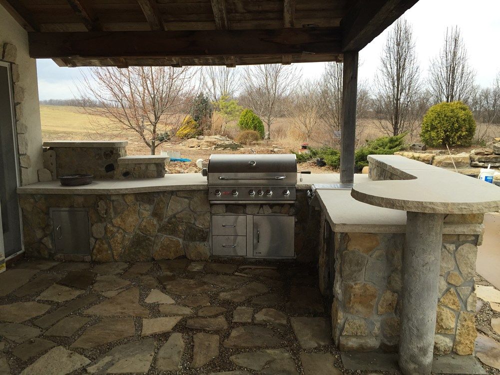 Outdoor kitchen with stone counters, grill, and bar under a wooden pergola on a flagstone patio.