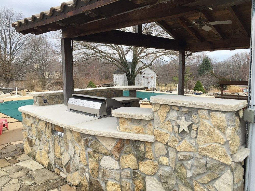 Outdoor kitchen with stone walls, grill, bar area under a wooden pergola, pool in background.