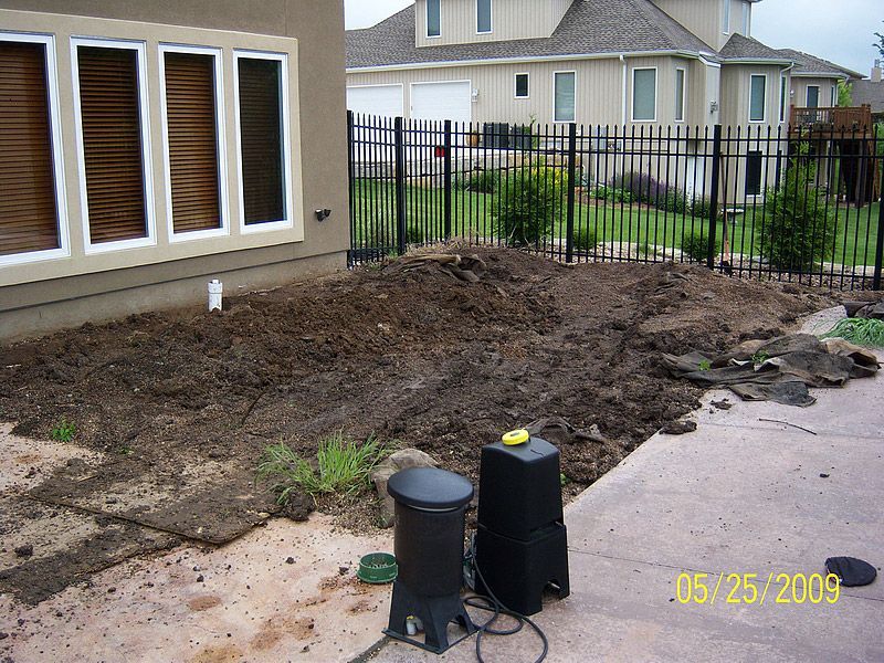 Yard with bare dirt, next to a patio and house.  Black speakers sit on the patio. A black fence surrounds the yard.