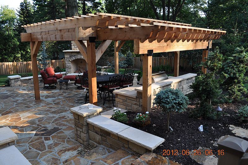 Wooden pergola over outdoor kitchen, stone patio, fireplace, and seating area.