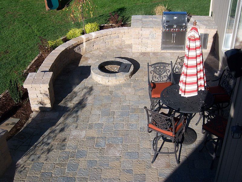 Patio with grill, fire pit, table and chairs; surrounded by landscaping and retaining wall.