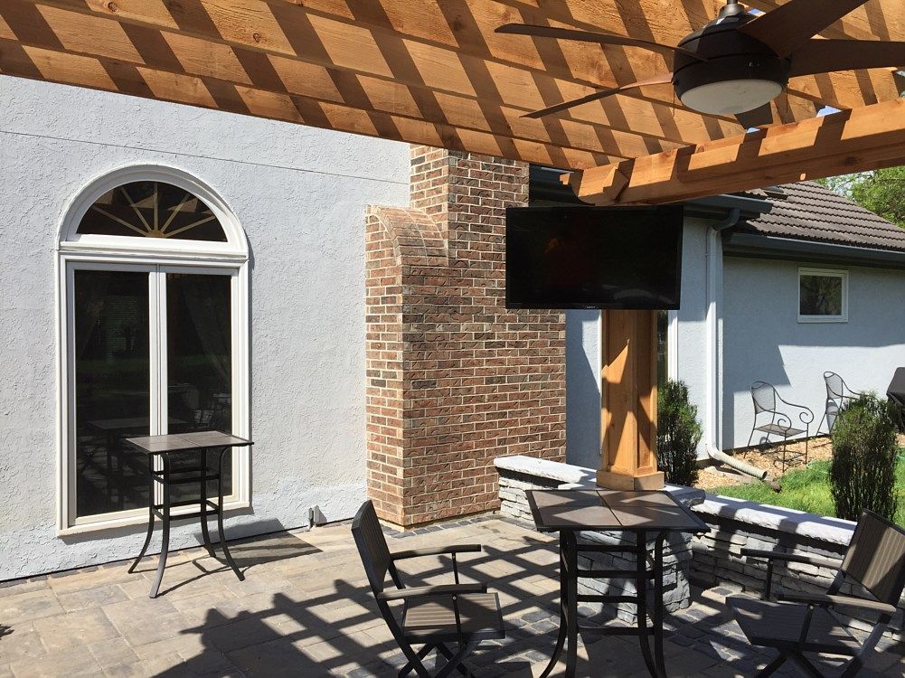 Patio with wooden pergola, brick chimney, and mounted TV. Tables and chairs on stone patio.