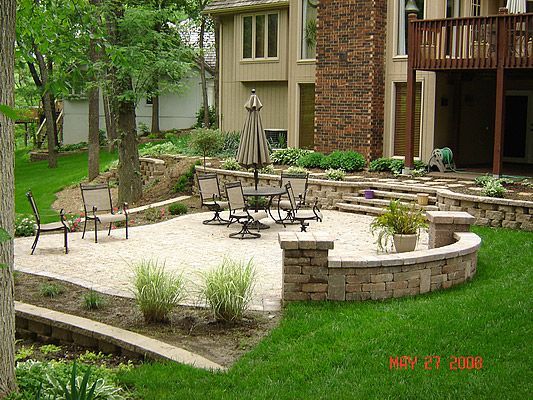 Stone patio with outdoor furniture surrounded by landscaping and a two-story house.