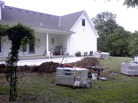 A backyard scene with house under construction, concrete patio, brick piles, and tools.