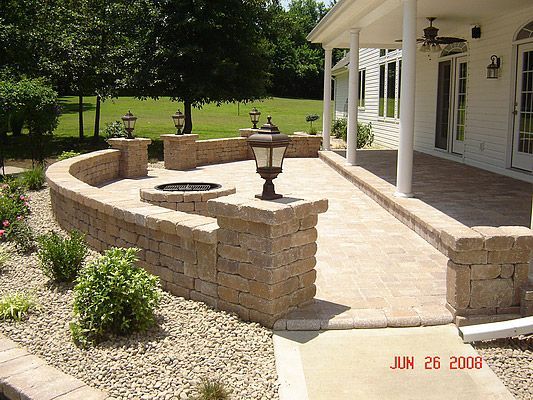 Brick patio with low walls, a fire pit, and lamp posts. Next to a white house with a porch.