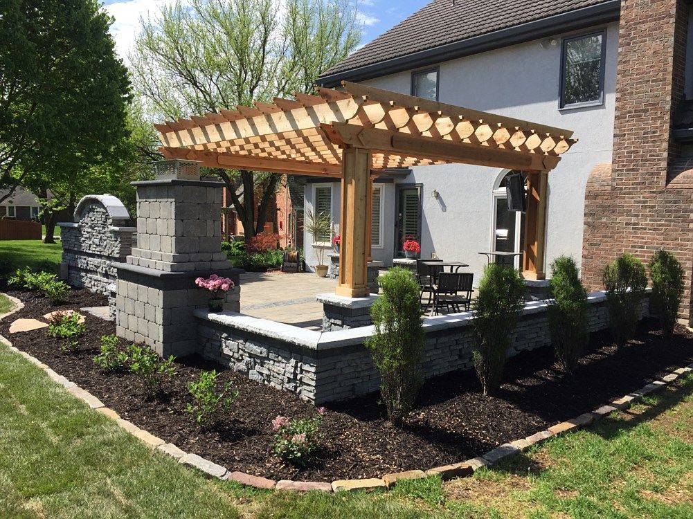 Patio with pergola, stone wall, and landscaping next to a house.