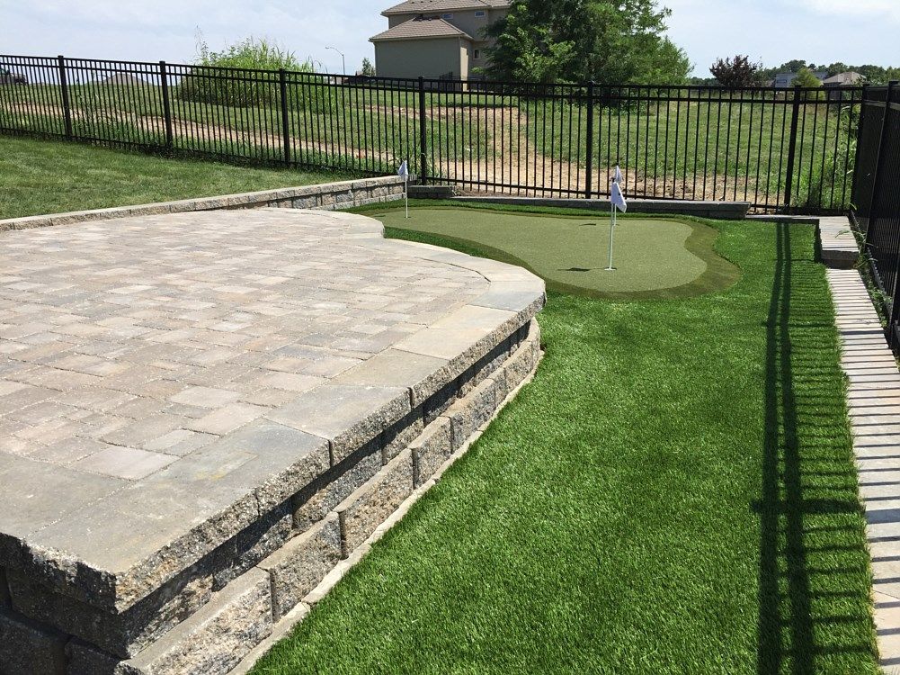 A backyard patio with a putting green, bordered by a retaining wall and black fence.