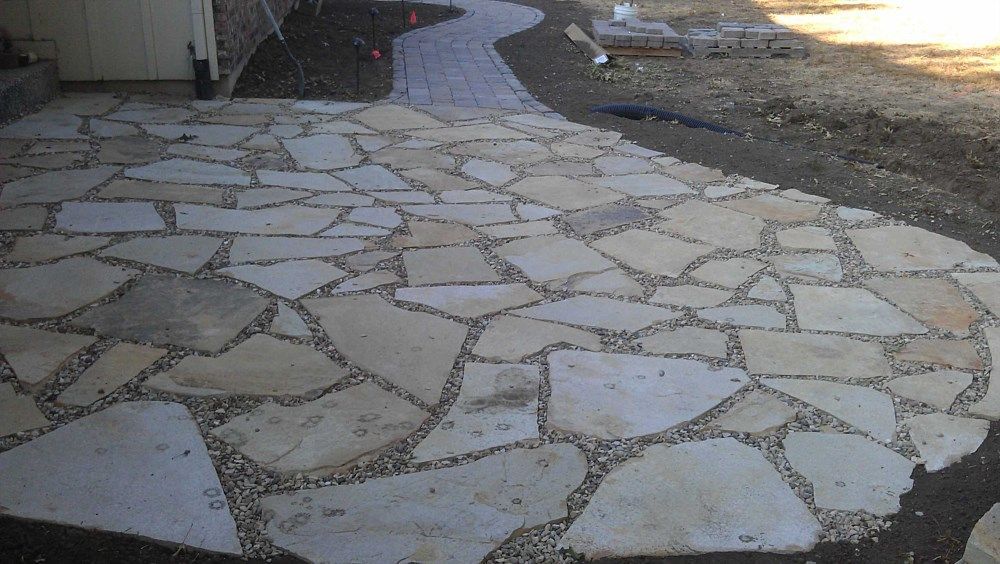 Stone patio with irregular-shaped pavers and small gravel filling spaces, next to a paved walkway.