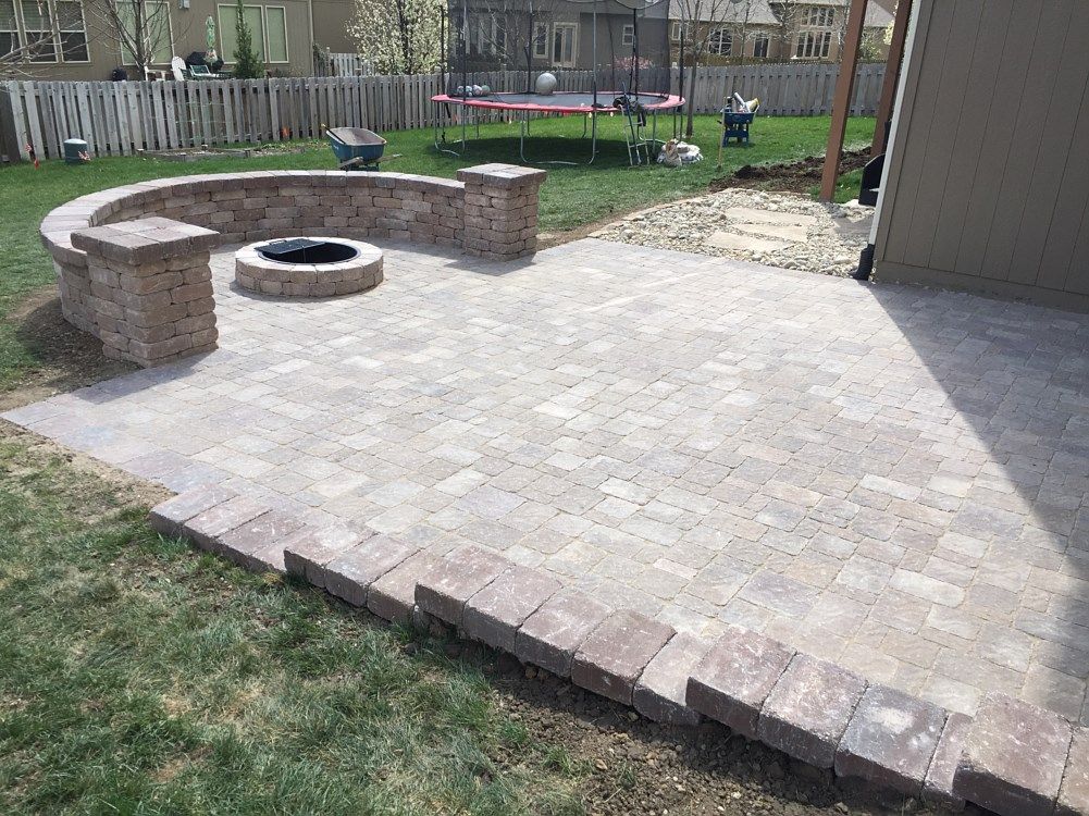 Brick patio with fire pit, bordered by low brick walls, adjacent to green grass and a house.
