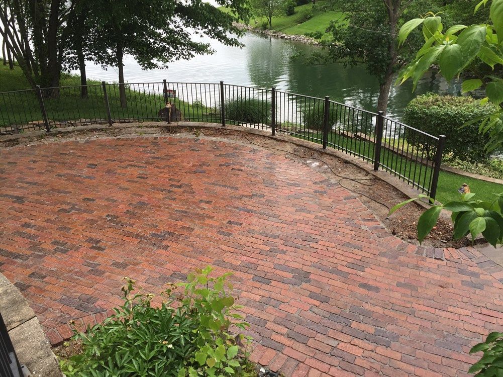 Brick patio with black fence overlooking a pond, surrounded by trees and greenery.