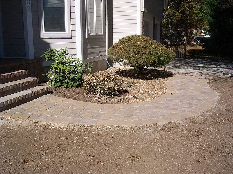 Brick walkway curving in front of house with landscaping and brown dirt.