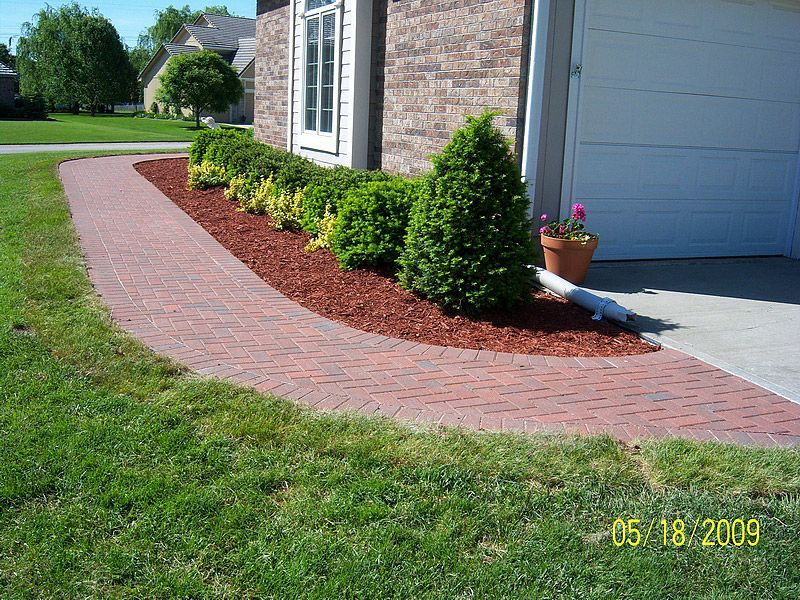 Brick pathway curves past a flower bed with shrubs, next to a garage and house with a green lawn.