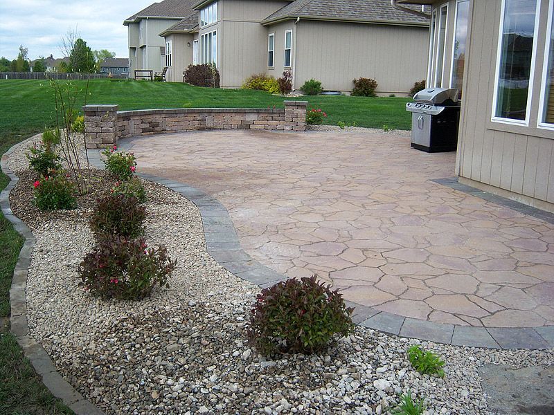 Patio with stone pavers, landscaping, brick wall, and a grill next to a beige house.
