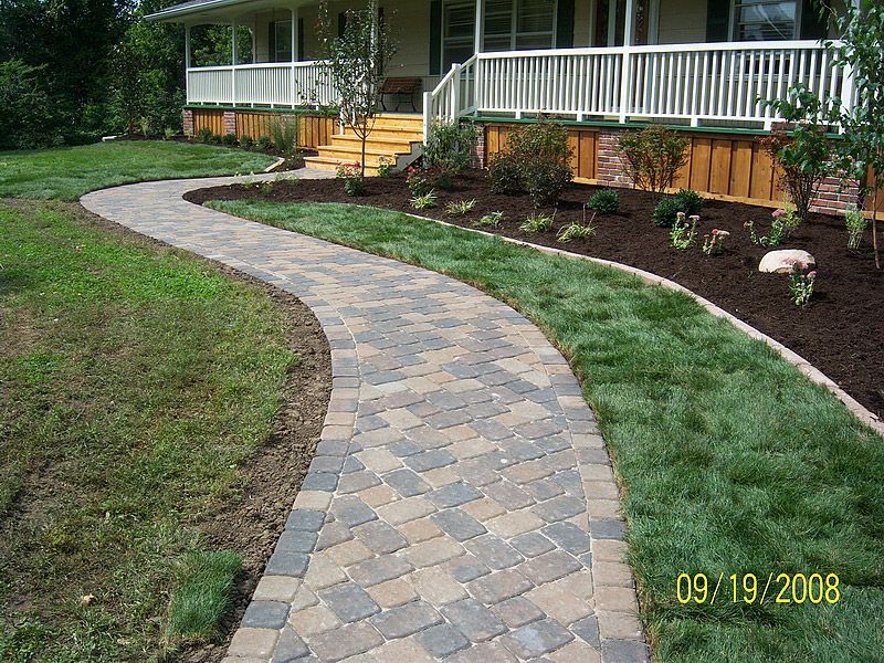 Brick pathway winding through green grass to a house with a white porch.