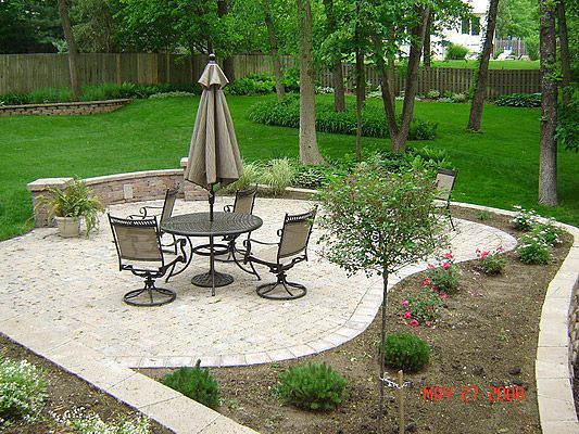 Patio with table and chairs, surrounded by flowerbeds, grass and trees in a backyard.