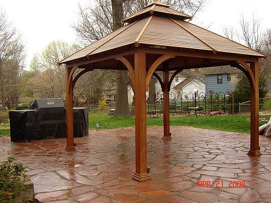 Wooden gazebo on a patterned stone patio, near a grill and a residential yard.