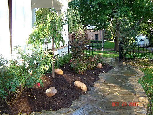 A stone pathway leads past a garden bed with plants, mulch, and large rocks toward a black gate.